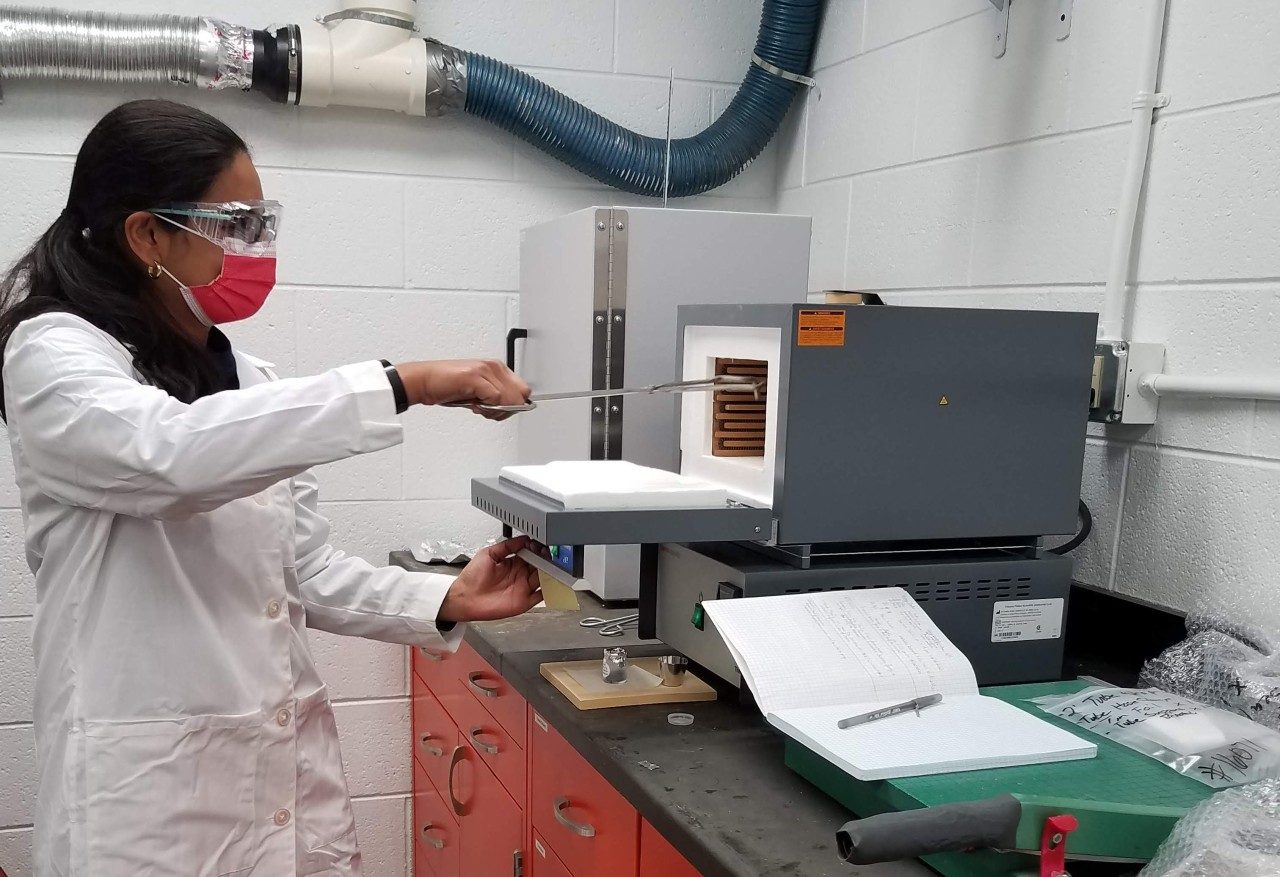 One person in a laboratory outfit (mask, safety glasses, and lab coat) putting a sample into a lab appliance
