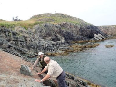 Two men crouch on a sloping rock with dramatic sea cliffs in the background.