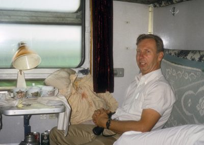 A man in his mid-50s, wearing a short-sleeved button-down shirt, sits comfortable inside a cluttered passenger train car.